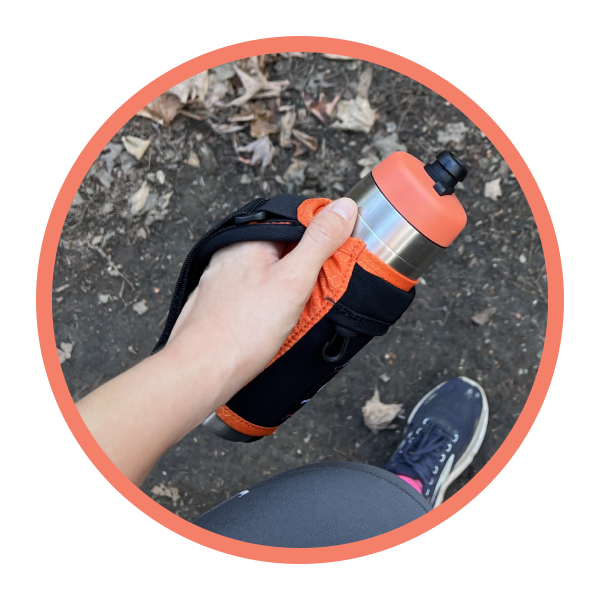 Hand holding a Bivo water bottle with an orange and black holder on a natural ground background
