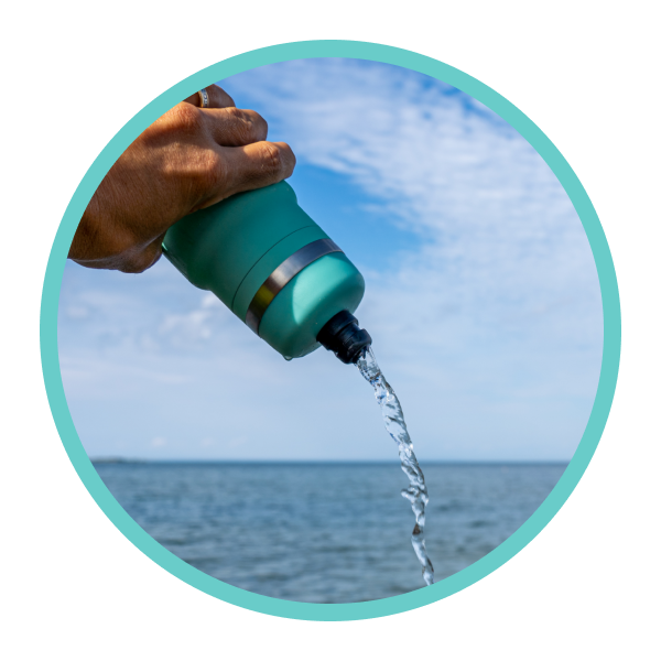 Hand pouring water from a Teal Bivo bottle into the ocean with a blue sky background