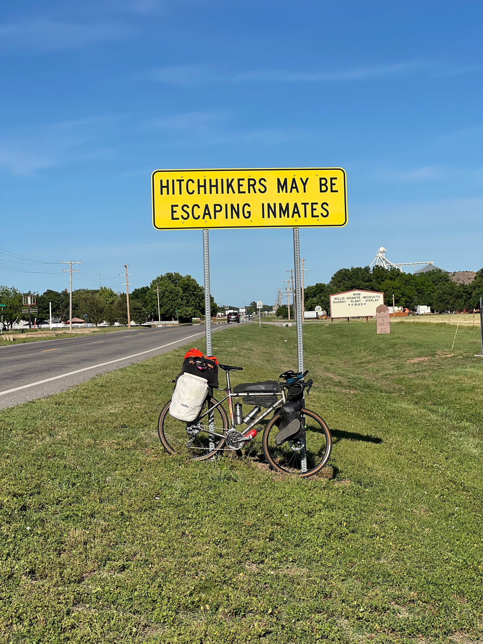 Bicycle with gear parked on grass next to a 'Hitchhikers May Be Escaping Inmates' sign on a road.