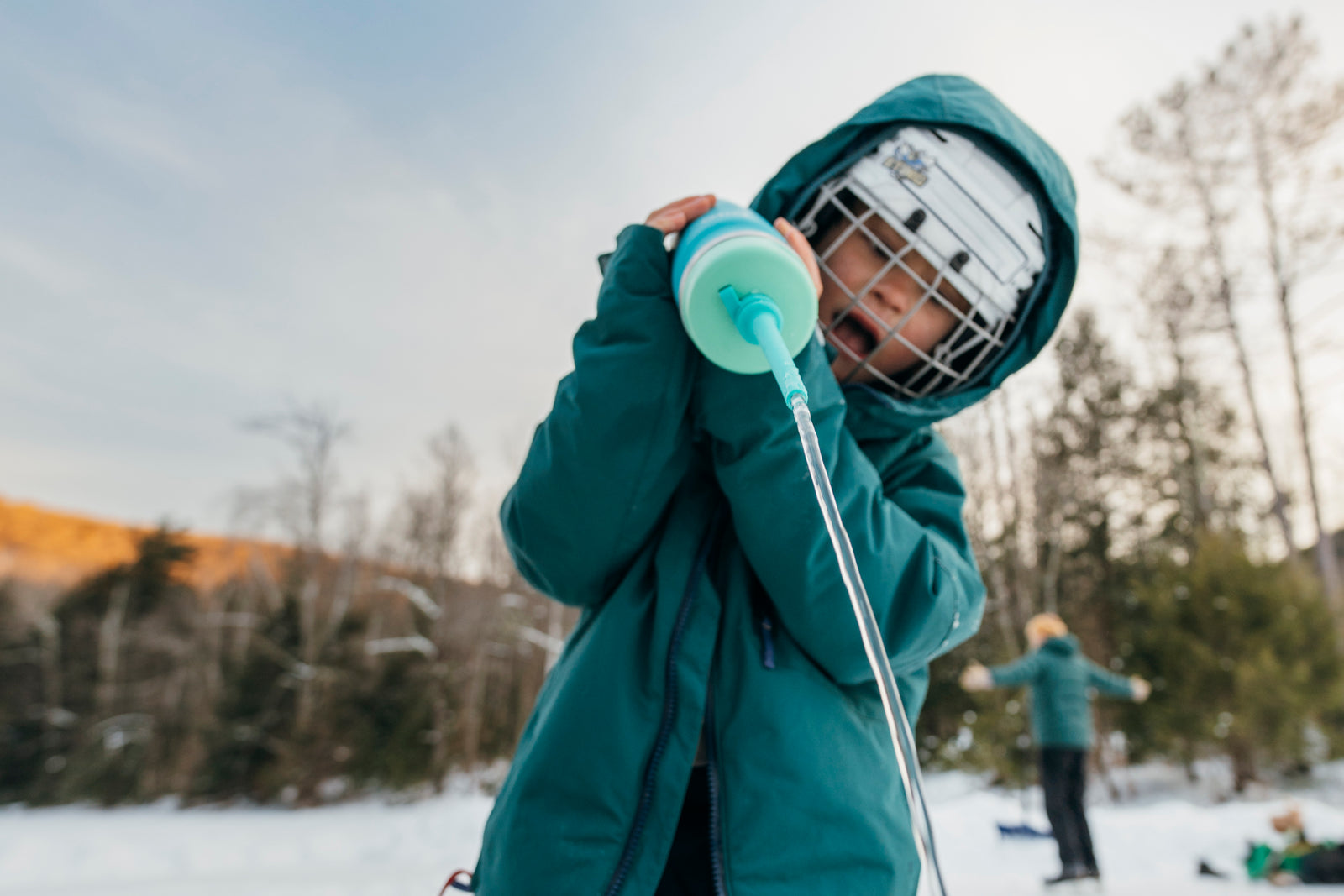 Child in teal jacket and helmet pouring water from a Bivo Gravity Flow Longshot Nozzle from a green bottle on a pond hockey rink in Vermont.