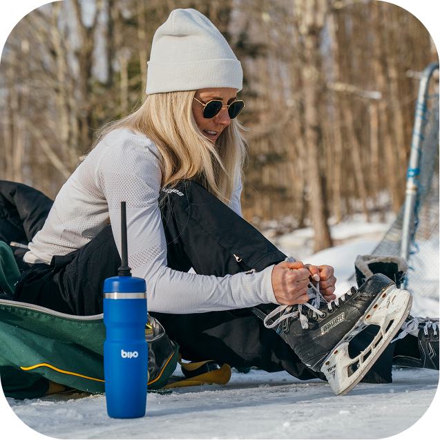 Woman preparing to ice skate in a snowy outdoor setting with a blue Bivo Hockey water bottle nearby.