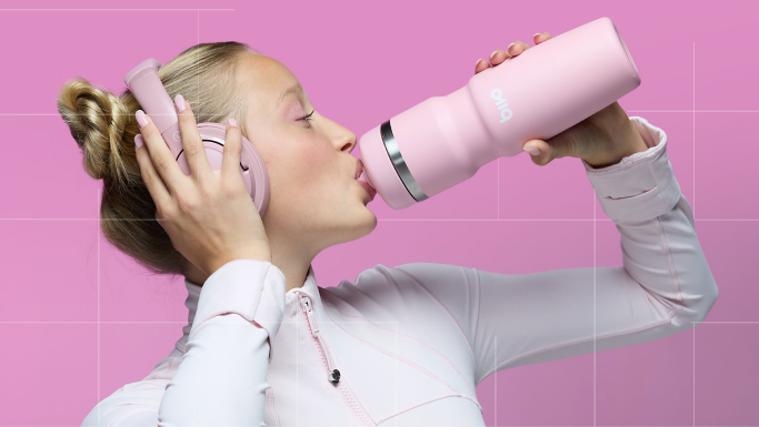 A woman in a white athletic zip-up top drinks from a light pink insulated bivo sports water bottle while wearing matching pink headphones against a pink grid background.