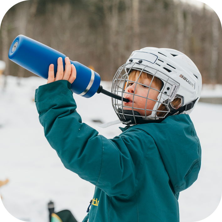Child in hockey gear and a mask holding a blue stainless steel water bottle for hockey outside, drinking easily