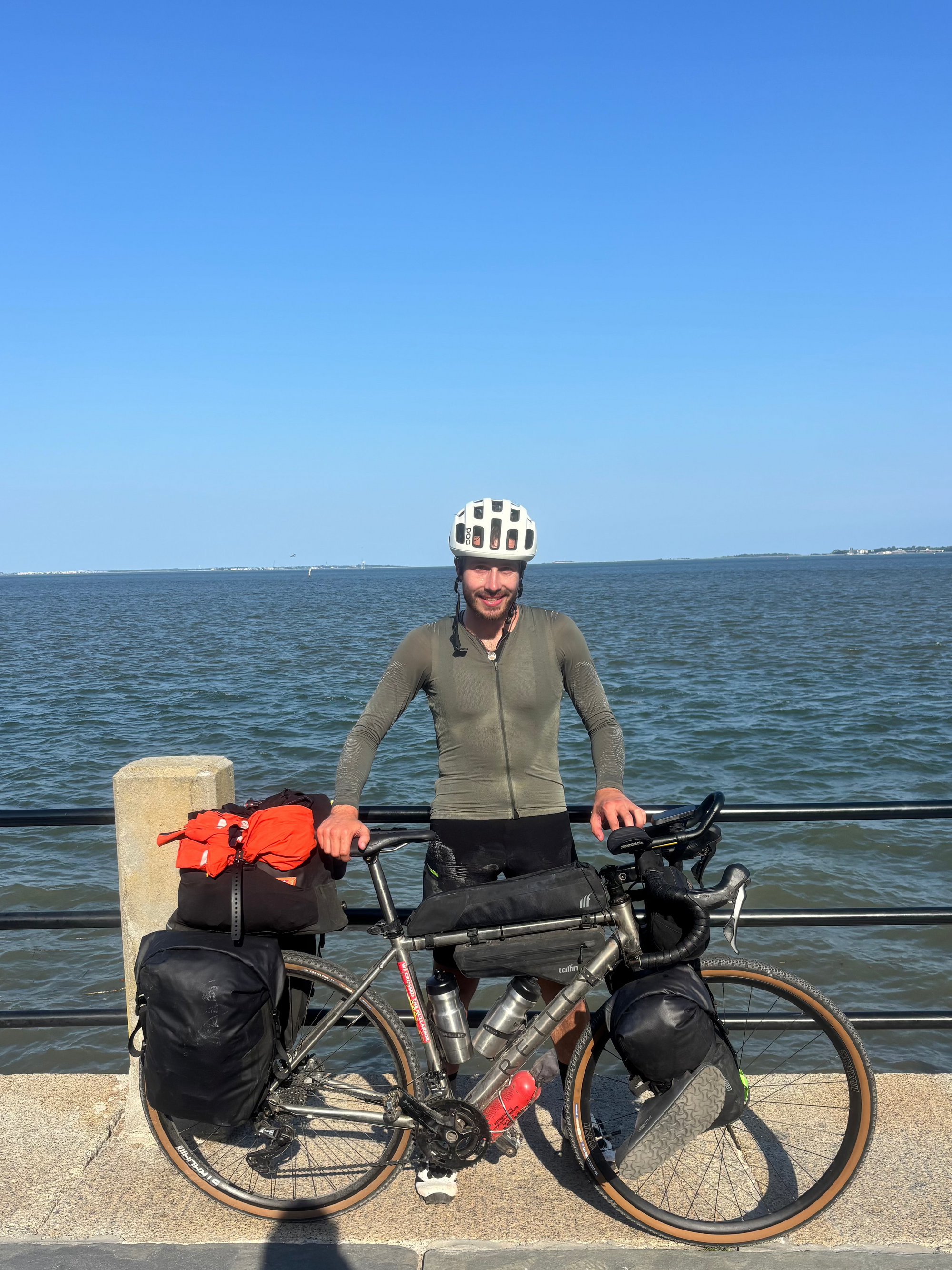 Man with a bicycle loaded with gear by a waterfront on a clear day